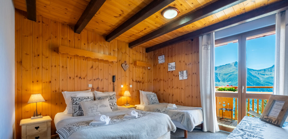 Chambre avec lit double et lit simple parés de plaids brodés, donnant sur un balcon montagne