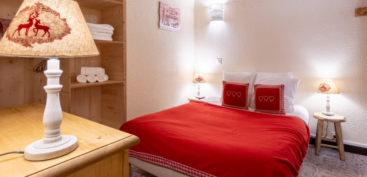 Lit double avec plaid rouge et coussins brodés de cœurs près d'une étagère en bois avec serviettes