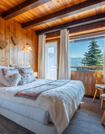 Lit double avec coussins brodés de cerfs et vue sur sapins et montagnes par la fenêtre