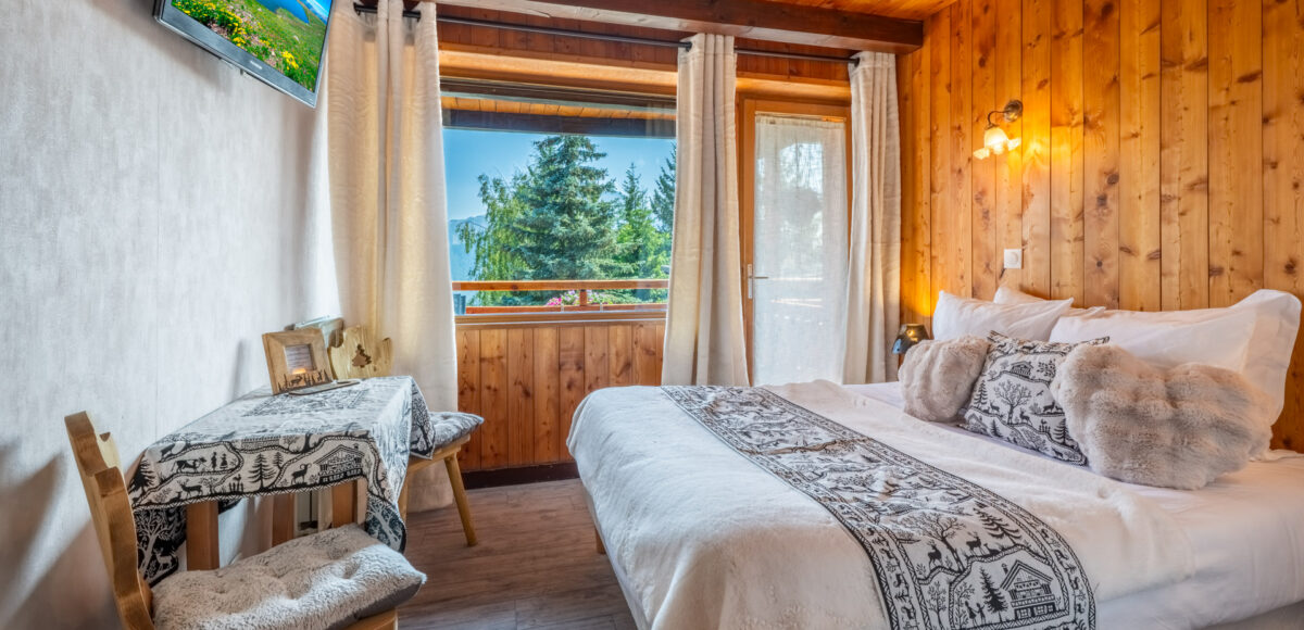 Lit double avec plaid en fourrure et coussins cœur, bureau en bois et vue sur les sapins